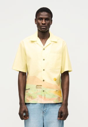 Άνδρας που φορά κίτρινο πουκάμισο με κοντά μανίκια και κουμπιά, με εκτύπωση τοπίου, συνδυασμένο με ανοιχτό μπλε τζιν, στέκεται μπροστά σε απλό φόντο.