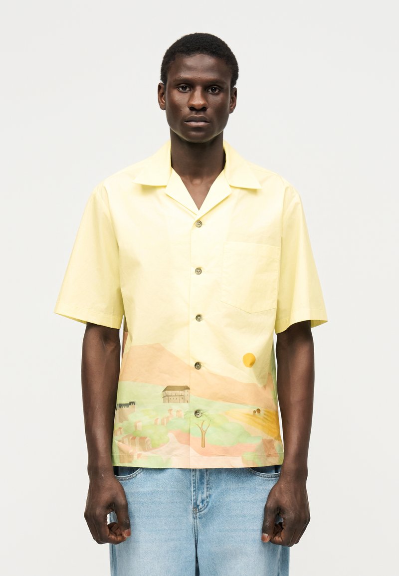 Άνδρας που φορά κίτρινο πουκάμισο με κοντά μανίκια και κουμπιά, με εκτύπωση τοπίου, συνδυασμένο με ανοιχτό μπλε τζιν, στέκεται μπροστά σε απλό φόντο.