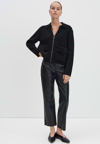 Vrouw staat met haar hand op de heup, gekleed in een zwarte geribbelde ritsjack, zwarte leren cropped broek en zwarte instap schoenen.