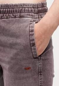 Δεξί χέρι τοποθετημένο στην τσέπη μοβ ξεβαμμένου τζιν με ελαστική μέση και μικρό κόκκινο κεντητό λογότυπο.