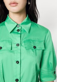 Grüne kurzärmlige Hemd mit Knopfleiste, Kragen, Brusttaschen und schwarzen Knöpfen. Glattes Material mit leichtem Glanz.