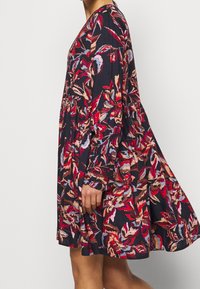Eine Frau trägt ein lockeres, langärmliges Kleid mit einem bunten Blumenmuster in Rot, Pink, Blau und Braun auf einem dunklen Hintergrund.
