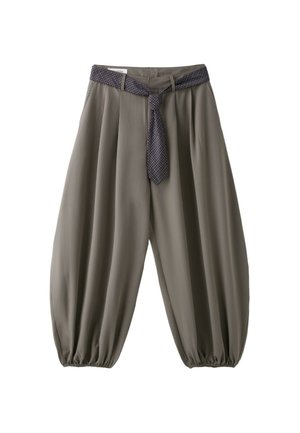Femme aux longs cheveux bouclés porte un haut blanc cintré à manches courtes, un large pantalon beige avec ceinture nouée et des talons blancs pointus sur fond gris.