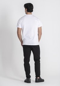 Biela bavlnená tričko, štandardný strih, krátke rukávy, v kombinácii s čiernymi džínsami a čiernymi teniskami, zobrazené zozadu na neutrálnom pozadí.