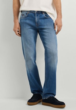 Mann trägt verblasste blaue Jeans mit geradem Bein, steht mit entspannten Händen, kombiniert mit schwarzen Schuhen und einem weißen, nicht in die Hose gesteckten Hemd.