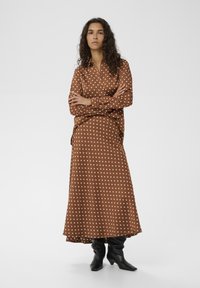 Brun polkaprikket bluse og matchende nederdel i et glat materiale, kombineret med sorte knæhøje støvler. Lange ærmer og løs pasform.