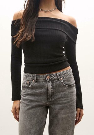 Vrouw draagt een zwarte off-shoulder geribde top met lange mouwen en hooggetailleerde vervaagde grijze jeans, staand tegen een effen achtergrond.