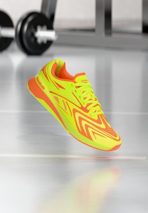 Chaussure de course Reebok jaune vif et orange flottant au-dessus d'un sol de salle de sport brillant avec des poids flous en arrière-plan.