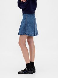 Jupe en denim, bleue, en forme de A avec un ourlet effiloché, portée avec des chaussures noires et des chaussettes. La tenue inclut un pull sombre, vue de côté.