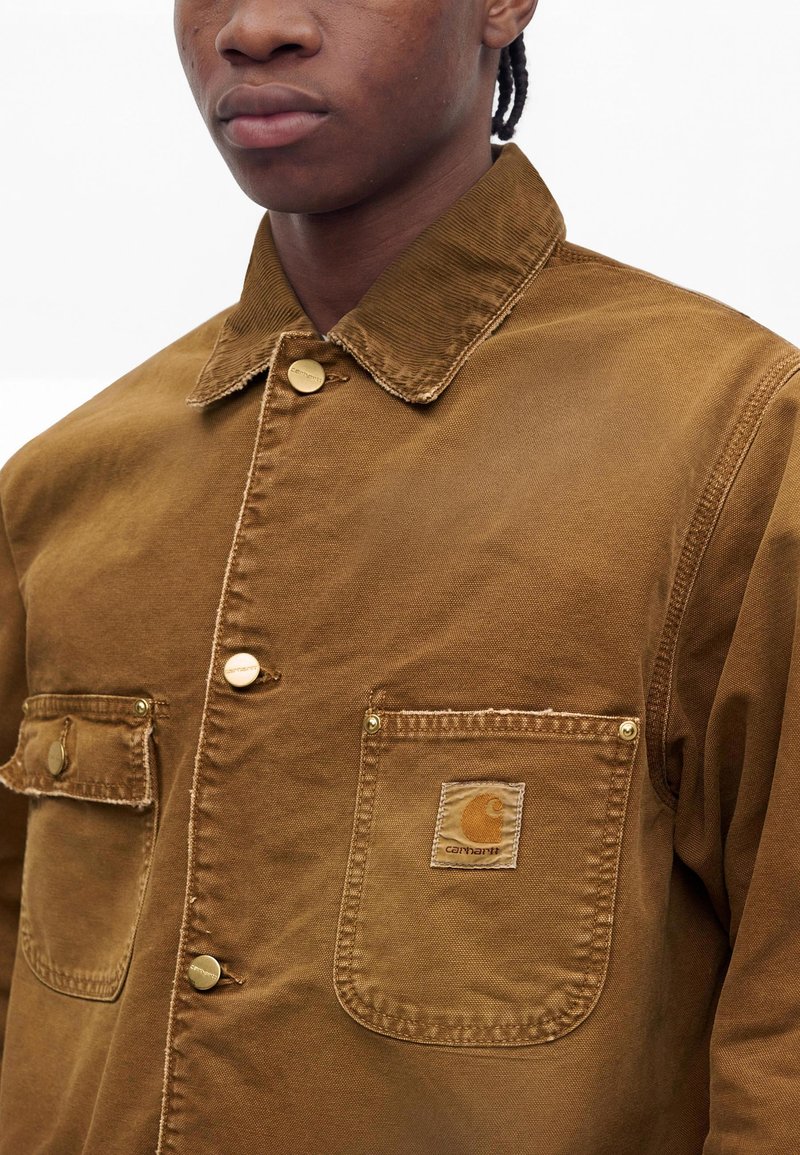 Uomo che indossa una giacca marrone Carhartt con bottoni, due tasche sul petto e colletto testurizzato, rivolto in avanti.