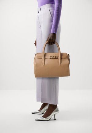 Borsa a mano in pelle beige con design strutturato, doppi manici e logo in rilievo. Abbinata a pantaloni viola chiaro e tacchi metallici argento.