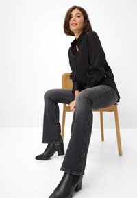 Chemise noire à boutons avec des manches bouffantes, associée à un jean évasé gris foncé et à des bottines à talons noires, assise sur une chaise en bois.