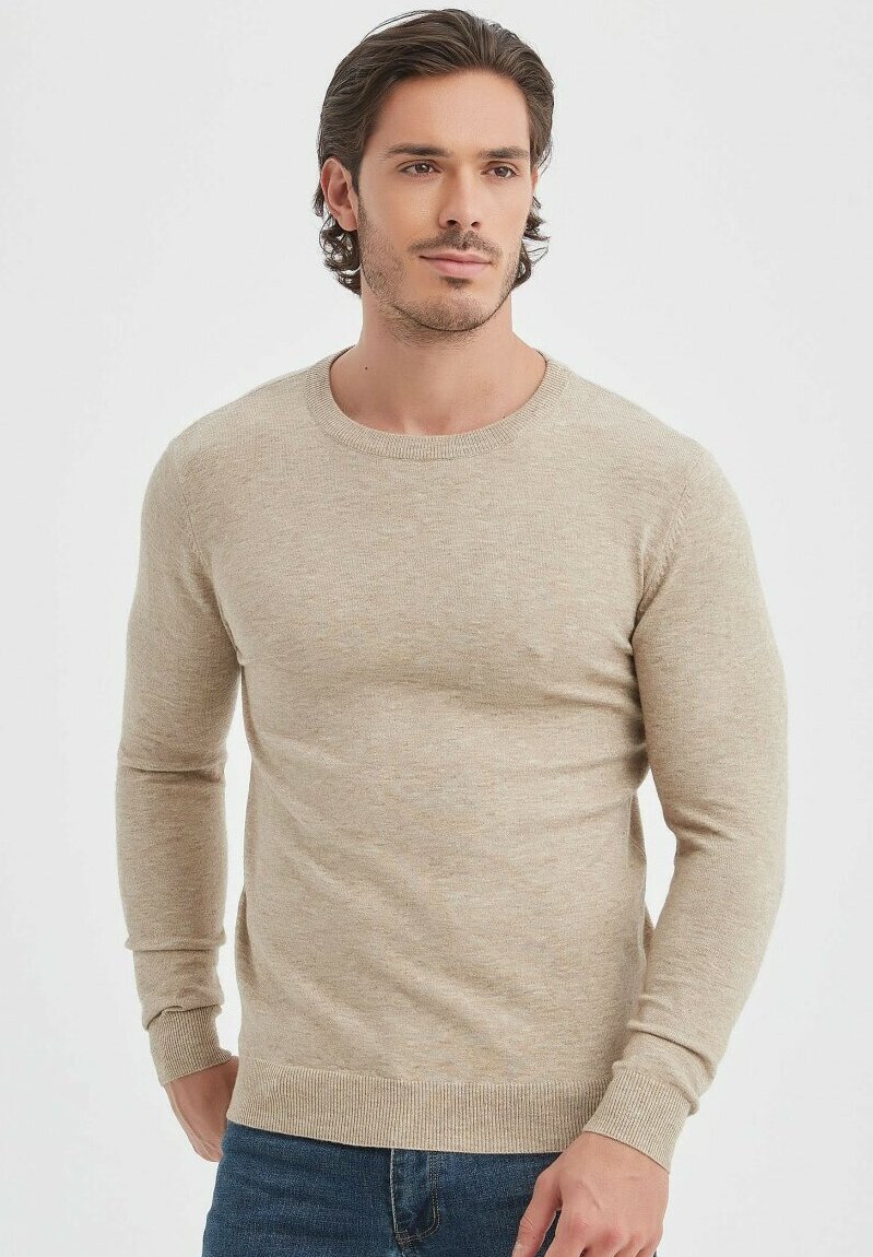 Pull à col rond beige à manches longues en tissu doux et texturé. Présente des poignets et un ourlet côtelés, porté par-dessus un jean en denim bleu.
