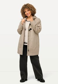 Cappotto beige con cappuccio e finitura testurizzata, dotato di chiusura con toggles, maniche lunghe e tasche laterali, abbinato a pantaloni neri a gamba larga.