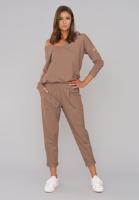 Brązowy sweter typu off-shoulder z długimi rękawami, zestawiony z dopasowanymi spodniami w kant z kieszeniami. Noszony z białymi sneakersami.