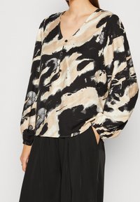 Sort og beige mønstret bluse med V-hals, knapdetaljer og lange, puffede ærmer. Lavet af letvægtigt stof med en glat tekstur.