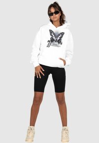 Haut à capuche blanc avec un graphisme de papillon et du texte, associé à un short noir. Porté avec des lunettes de soleil et des baskets de couleur claire.