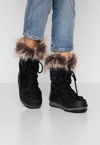 Person som bär svarta vinterkängor med pälsdetalj och ljusblå uppvikta jeans, stående på ett vitt golv.