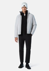 Hellgraue Steppjacke mit hohem Kragen, darüber ein schwarzer gerippter Pullover. Schwarze Hose und schwarze Turnschuhe vervollständigen den Look. Weiße Mütze auf dem Kopf.