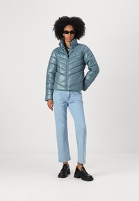Tirkīza krāsas pūkains mētelis ar augstu apkakli un horizontālām šuvēm, apvienots ar gaiši ziliem džinsu biksēm un melnajām loafers. Modelis valkā saulesbrilles.