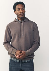 Felpa marrone testurizzata con cappuccio regolabile, indossata sopra una camicia a quadri, abbinata a jeans scuri, che mostra una vestibilità comoda e un motivo unico.