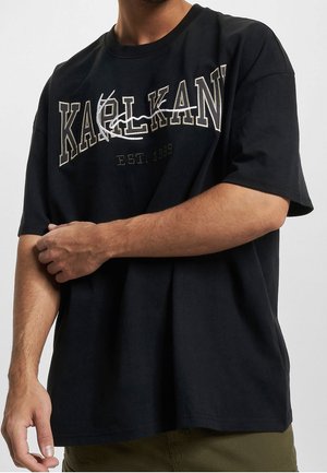 Mann trägt schwarzes oversized T-Shirt mit dem Text „KARL KANI EST. 1989“ und weißem grafischem Design auf der Vorderseite, kombiniert mit olivgrüner Hose.