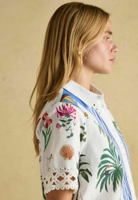 White shirt with colorful floral and tropical prints, featuring blue accents and lace trim on the sleeves. Collar and button fastenings visible.
