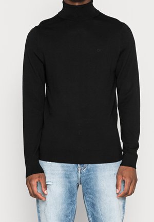 Homme portant un pull à col roulé noir et un jean bleu clair déchiré, debout devant un fond blanc uni.