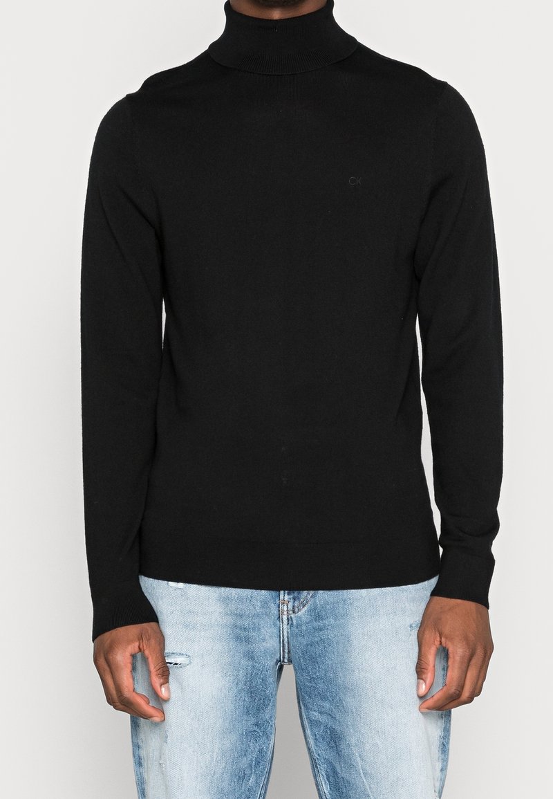 Homme portant un pull à col roulé noir et un jean bleu clair déchiré, debout devant un fond blanc uni.