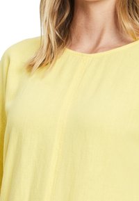 Żółta, lekka bluzka z okrągłym dekoltem i miękką teksturą. Posiada subtelne detale w postaci przeszyć z przodu.