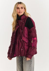 Bordová prešívaná bunda s textúrovaným čiernym ramenom, patentkami a kvetinovým akcentom na jednej strane. Zahrňuje bočné vrecká.