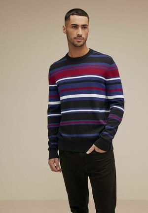 Zwarte trui met horizontale strepen in rood, blauw en wit, ronde hals, ribgebreide manchetten en zoom, gedragen met zwarte broeken.