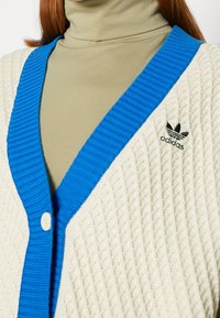Stickad cardigan i kr ämfärg med ett strukturerat mönster, som har en ribbad blå V-halsringning och en vit knapp; inkluderar svart Adidas-logotyp. 