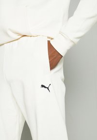 Hvid hættejakke med kengurulomme og joggers med elastisk talje. Har et sort Puma-logo broderet på benet. Glat tekstur.