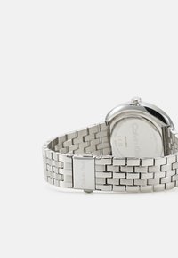 Sølv rustfritt stålklokke med et polert rundt hus, rutenettmønsteret armbånd og gravert logo på spennen. Baksiden er glatt og sirkulær.