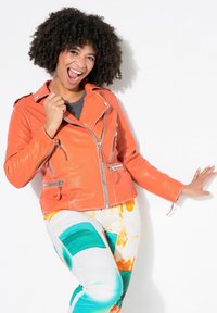 Orangene Leder-Motorradjacke mit mehreren Reißverschlüssen und Taschen, kombiniert mit mehrfarbigen gemusterten Hosen mit blauen und orangenen Akzenten.