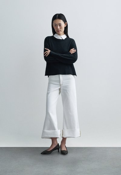 Pull noir en laine tricotée avec ourlet côtelé, superposé à une chemise blanche à col. Pantalon blanc à jambes larges avec revers effilochés, assorti à des talons noirs.