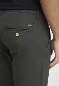 Grijze broek van gladde stof, met een achterzak met knoopsluiting en een klein label voor branding.