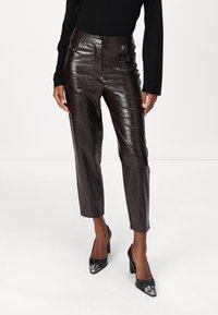 Braune Kunstlederhosen mit gemustertem Krokodilleder, hoher Taille, verkürzter Länge und Schlitzdetails am Saum, kombiniert mit schwarzen High Heels.