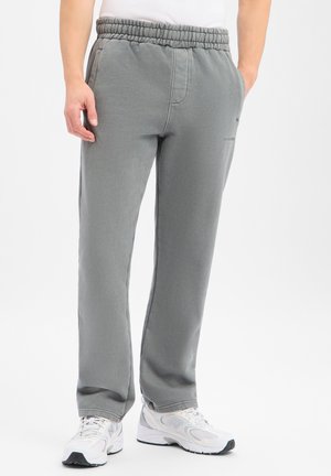Homme portant un pantalon de survêtement gris ample avec une taille élastique et des chaussures de sport blanches, debout devant un fond blanc uni.