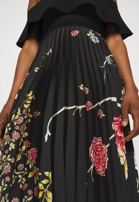 Faltenkleid mit schwarzem Stoff-Untergrund, das lebendige Blumenmuster in Rot, Blau und Gelb zeigt. Rüschenhalslinie mit strukturierten Falten.