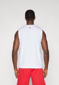 Άσπρο βαμβακερό φανελάκι χωρίς μανίκια με μικρό κεντητό λογότυπο στο πίσω μέρος, συνδυασμένο με κόκκινα σορτς. Απλός σχεδιασμός και άνετη εφαρμογή.