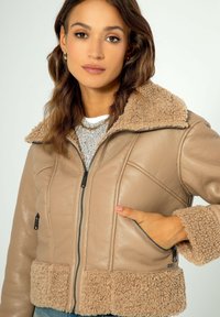 Giacca di pelle color tan con colletto e polsini beige pelosi. Presenta una chiusura a zip, due tasche frontali e dettagli testurizzati sulle maniche.
