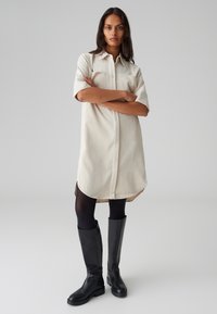 Abito camicia beige con colletto, maniche corte, spacchi laterali e silhouette dritta, abbinato a stivali in pelle nera al ginocchio.