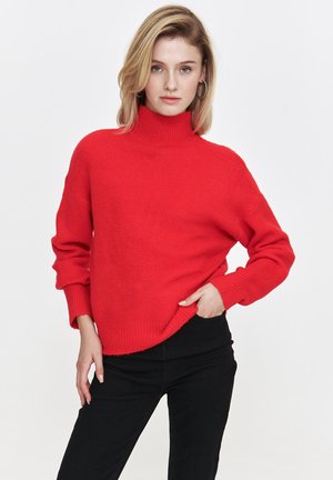 Czerwony sweter z golfem, dziergany, o luźnym kroju, z prążkowanymi mankietami i dołem. Noszony z czarnymi dopasowanymi spodniami, o gładkiej fakturze.