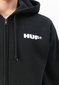 Svart zip-up hettegenser med hette og snøring, med hvit "HUF®" logo brodert på venstre bryst.
