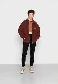 Casaco de bombazina castanho com gola, com um patch de logótipo, usado por cima de um top curto às riscas, jeans skinny pretos e ténis castanhos de cano alto.