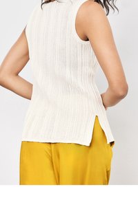 Biała, bezrękawnikowa bluzka z prążkowanego dzianiny z bocznym rozcięciem, noszona z jaskrawymi żółtymi satynowymi spodniami, sfotografowana z tyłu na wysokości talii.