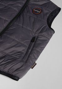 Gilet rembourré gris avec fermeture éclair à l'avant, un écusson logo sur la poitrine gauche, et une poche zippée supplémentaire sur le côté. Texture lisse et léger.