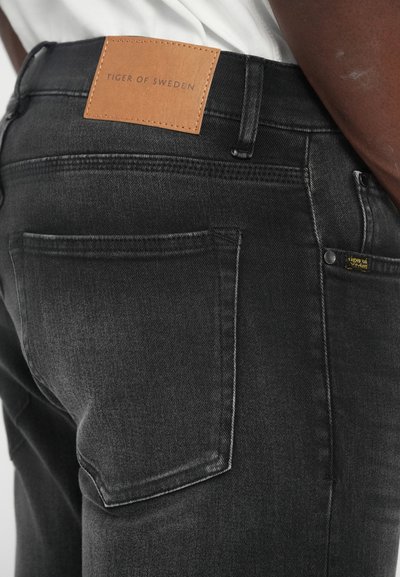 Μαύρα τζιν παντελόνια από денim με δερμάτινο λογότυπο που λέει "TIGER OF SWEDEN." Χαρακτηριστικά περιλαμβάνουν πέντε τσέπες και αντίθετη ραφή.